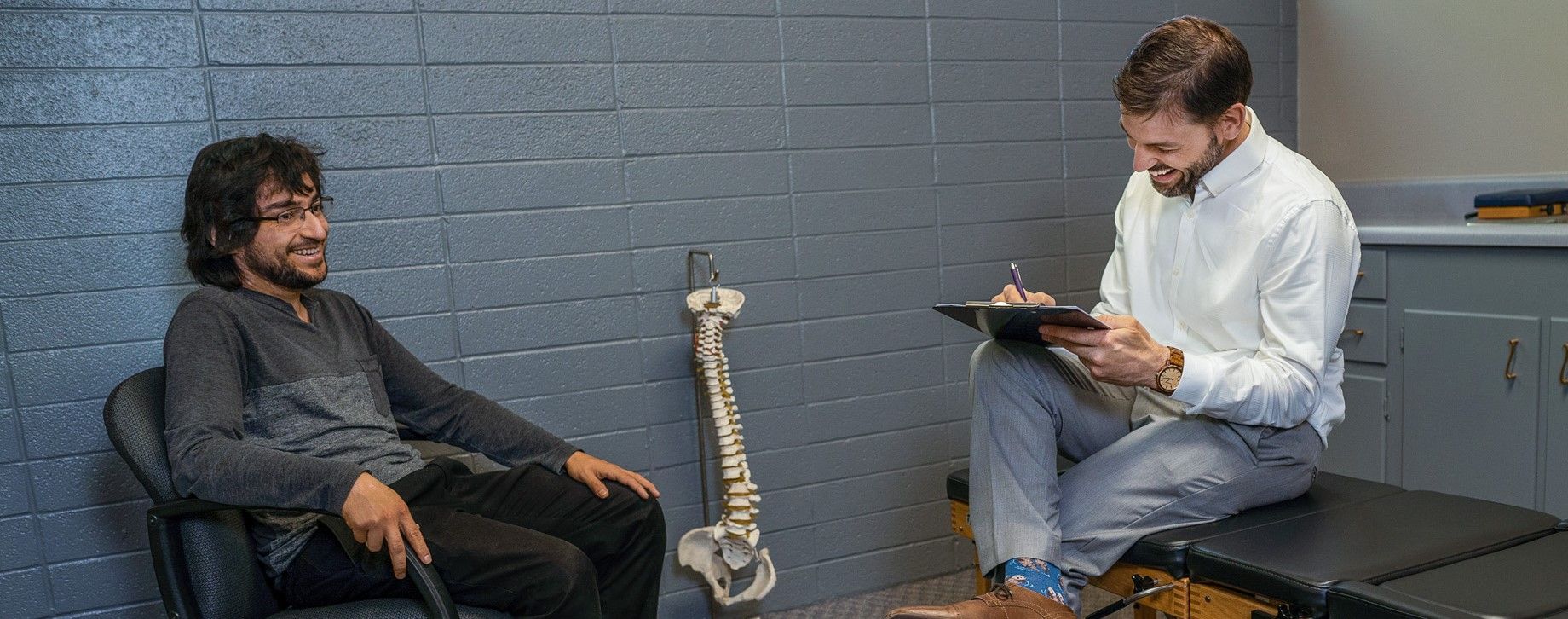 Chiropractor smiling while filling out patient note while patient sits in chair relaxed smiling