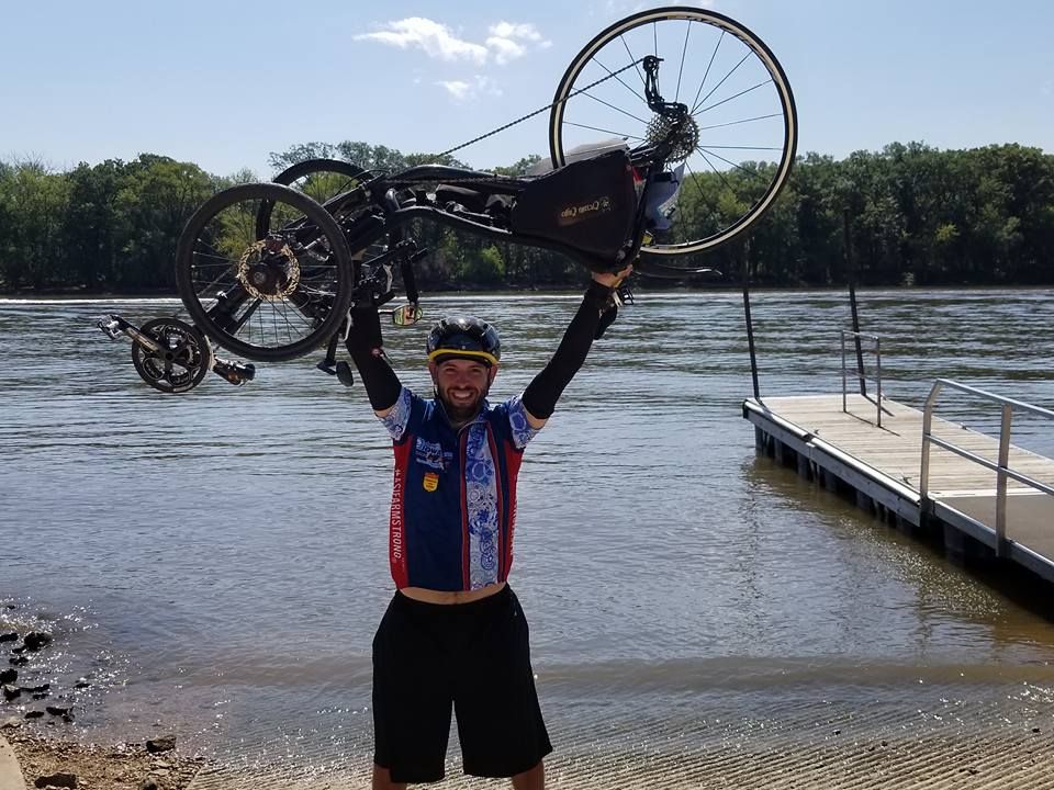 Dr. Clint Mausser holding his bicycle overhead beside a river